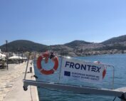 A banner and two lifebuoys are attached to a retractable bridge of a ship. The banner says Frontex European Border and Coast Guard Agency. In the background there is water, part of the promenade and the town of Vathy.